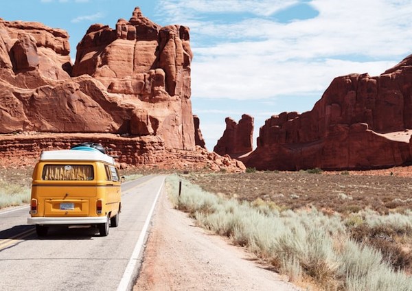 a yellow van on a road with a large rock formation in the background with Arches National Park in the background